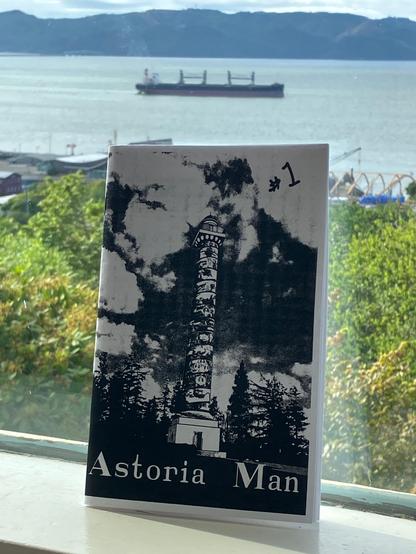 Cover of my zine "Astoria Man", featuring black and white depiction of the Astoria Column.  Zine is leaning up against a window behind which is a view of a bulk carrier ship on Columbia River in Astoria, Oregon and the mountains across the river in Washington.