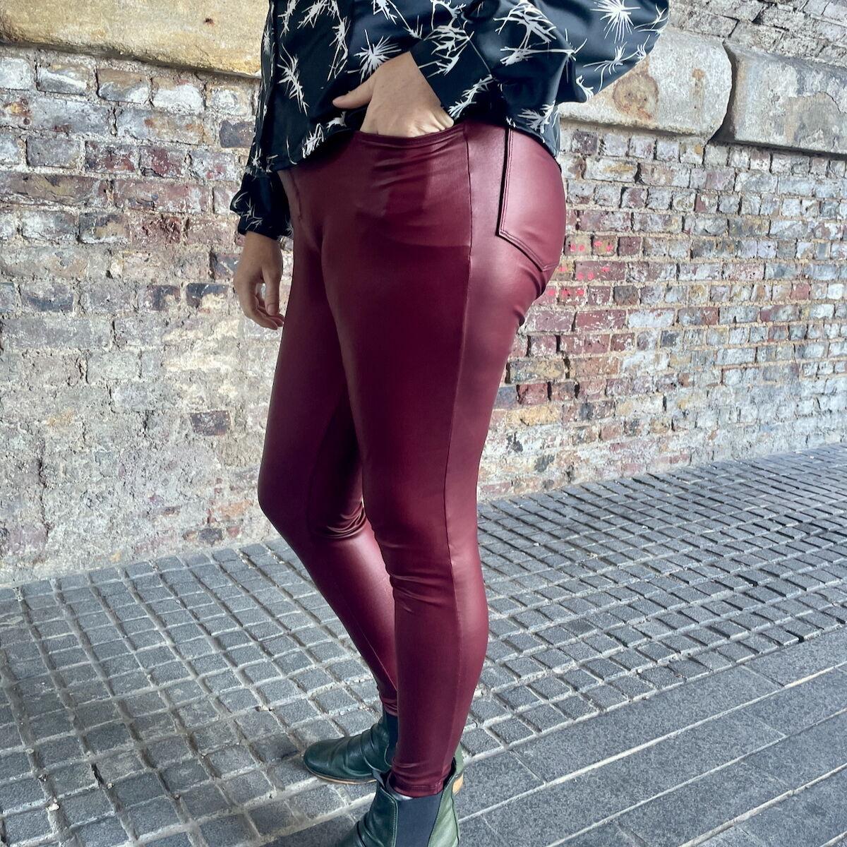 Detail of a woman wearing tight-fitting, burgundy pleather trousers with jeans-style pockets front and back. She's standing outside with a brick wall behind.
