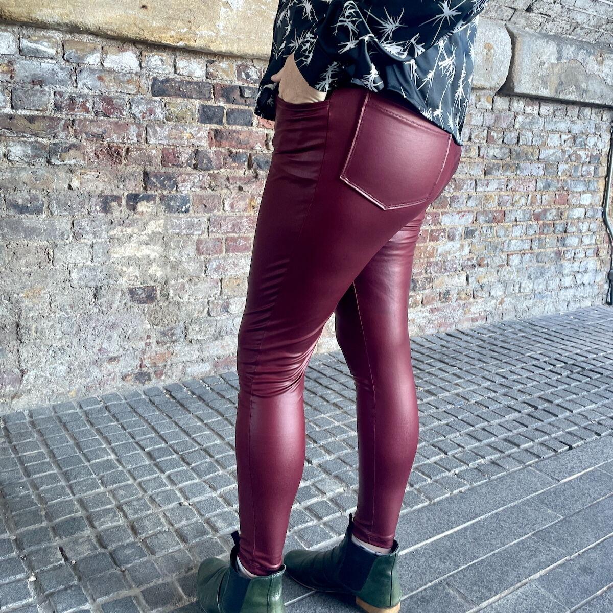 Detail of a woman wearing tight-fitting, burgundy pleather trousers with jeans-style pockets front and back. She's standing outside with a brick wall behind.