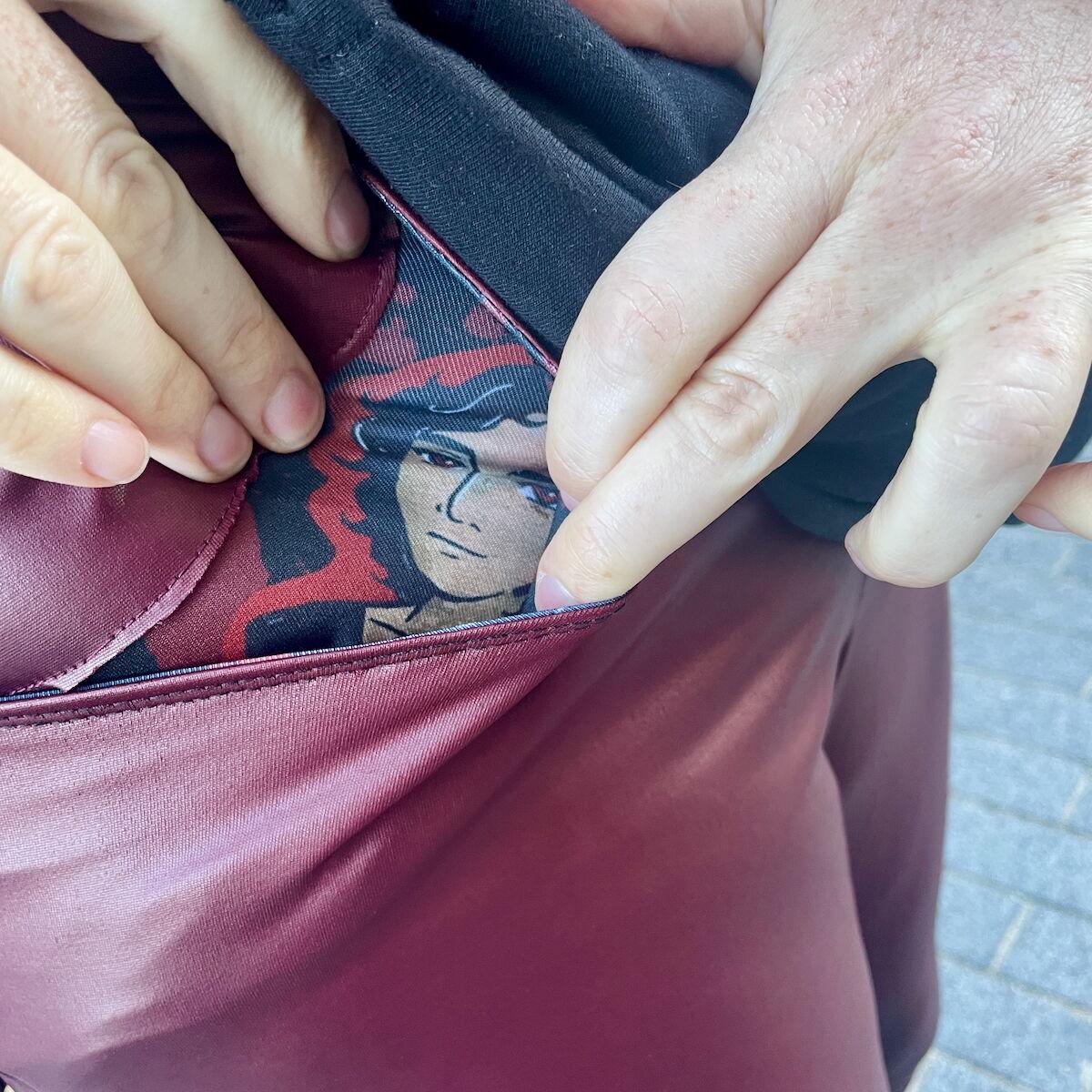 Detail of white hands reaching into the front pocket of a pair of dark red pleather trousers to reveal the pocket lining fabric featuring an illustration of Armand.