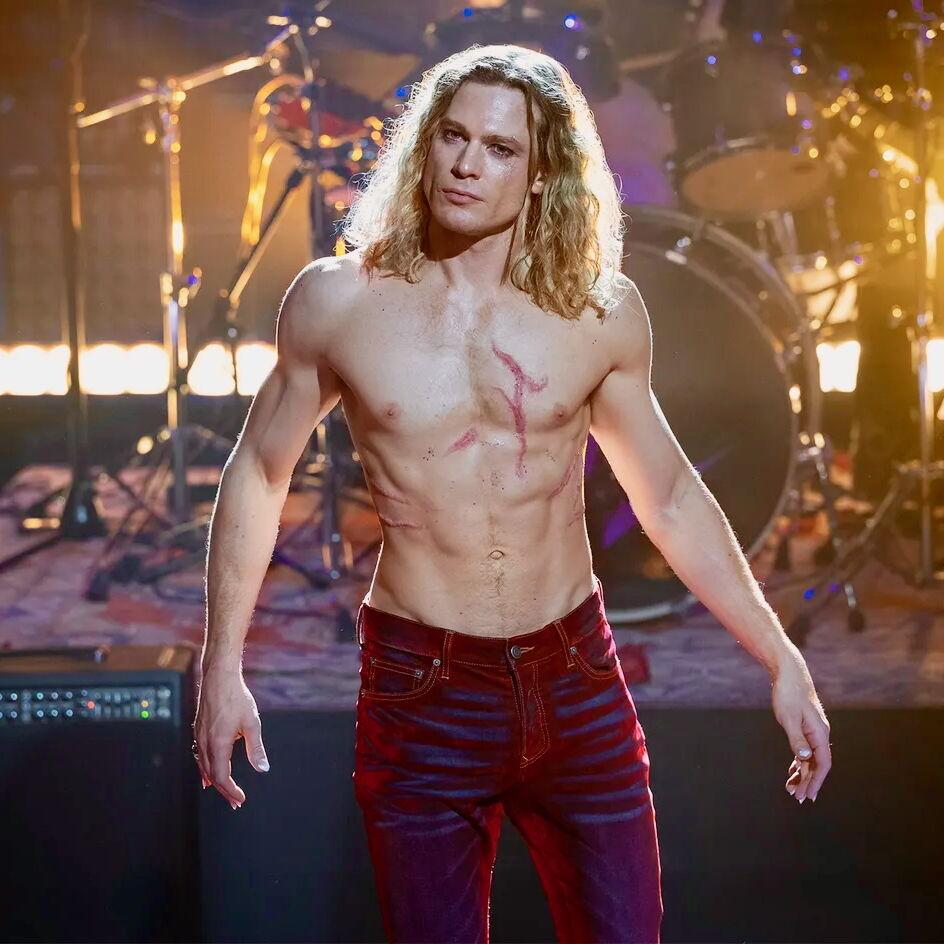Sam Reid as Lestat is standing shirtless on stage in front of a drumkit, wearing low-slung dark red trousers. His chest is scarred and he's looking bewildered.