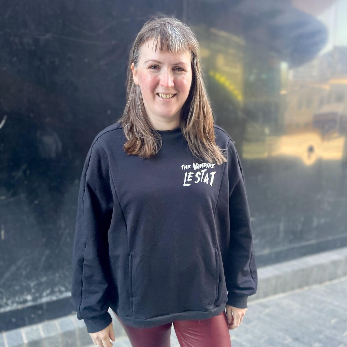 A white woman with brown hair is standing outside wearing an oversized black sweatshirt with "The Vampire Lestat" scrawled on the chest in white vinyl.