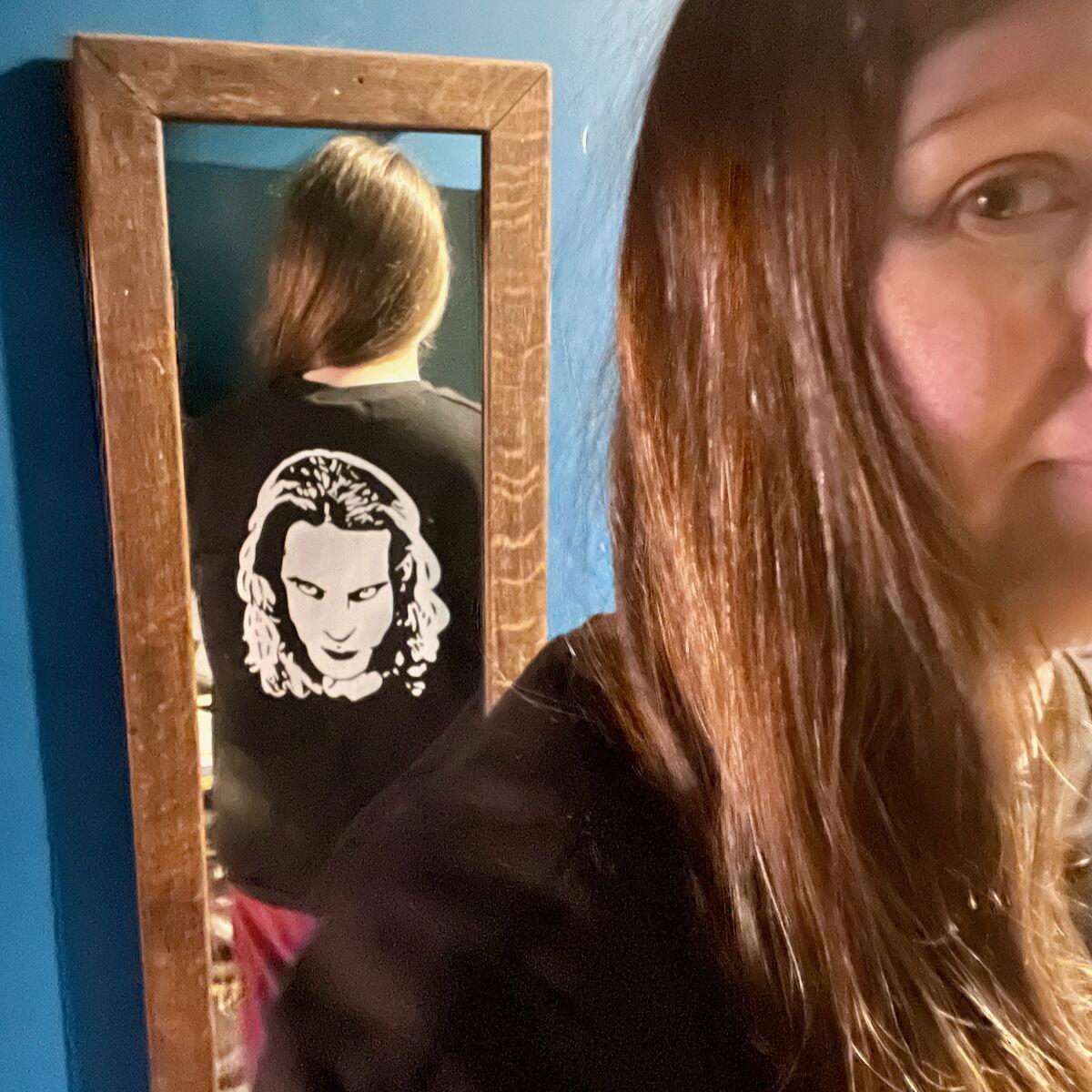A white woman with long brown hair is taking a selfie with her face partially seen, but her back is visible in a mirror. She’s wearing a black sweatshirt with a looming man’s face in white vinyl on the back.