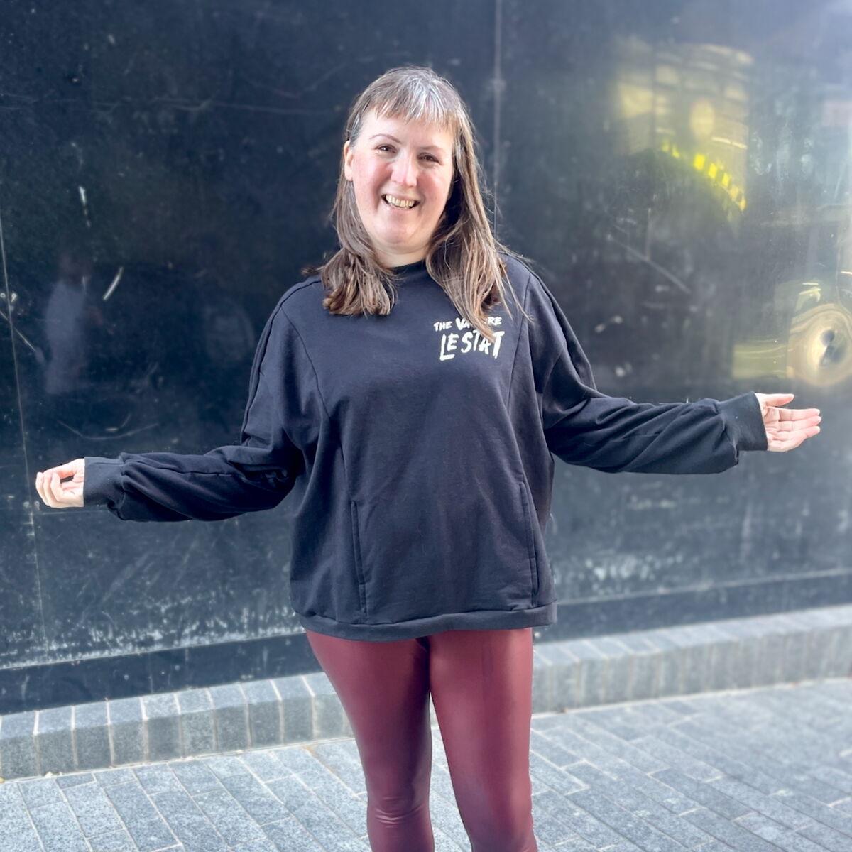 A white woman with brown hair is standing outside wearing an oversized black sweatshirt with "The Vampire Lestat" scrawled on the chest in white vinyl. Her arms are outstretched and she's smiling.