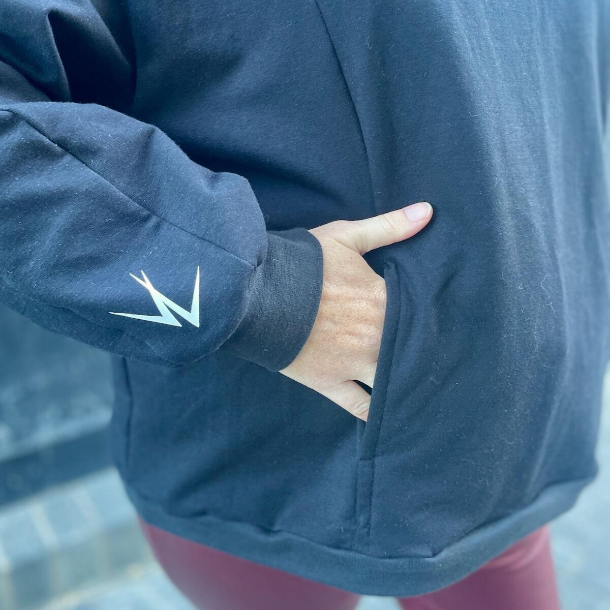 Detail of the kangaroo pocket on an oversized black sweatshirt. A white hand is partially in the pocket and a white, interlocking VL symbol is on the sleeve.