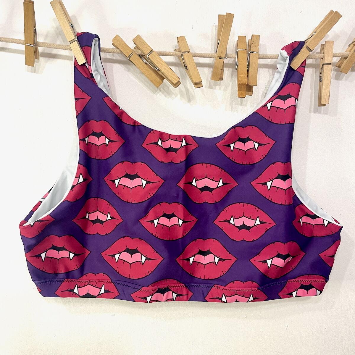 A sports bra made of purple lycra with red fanged lips is hanging on a clothes line against a white wall.