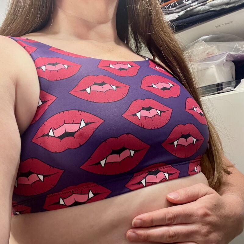A white woman is wearing a sports bra made of purple lycra with red fanged lips and her face cannot be seen.