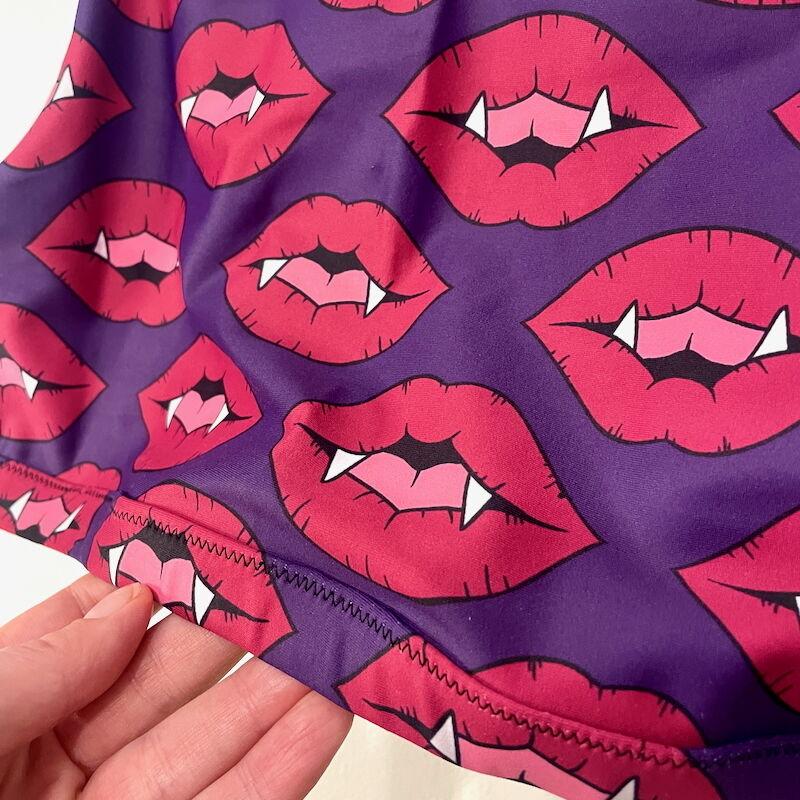 A white hand holds a sports bra made of purple lycra with red fanged lips. A portion of the garment can be seen, including the zigzag topstitching on the elastic bottom band.