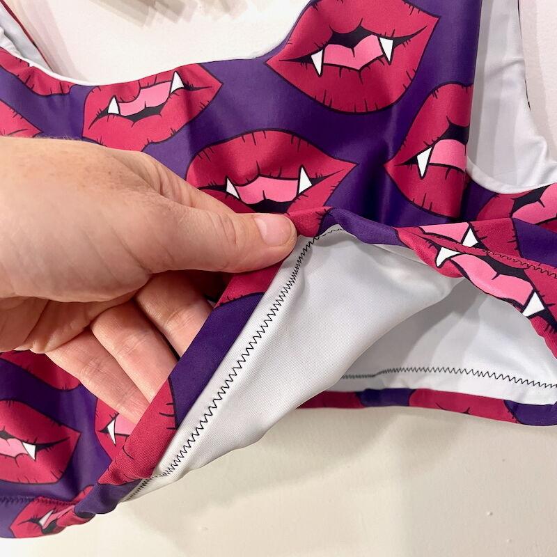 A white hand flips up the bottom of a sports bra made of purple lycra with red fanged lips so that the white lining can be seen.