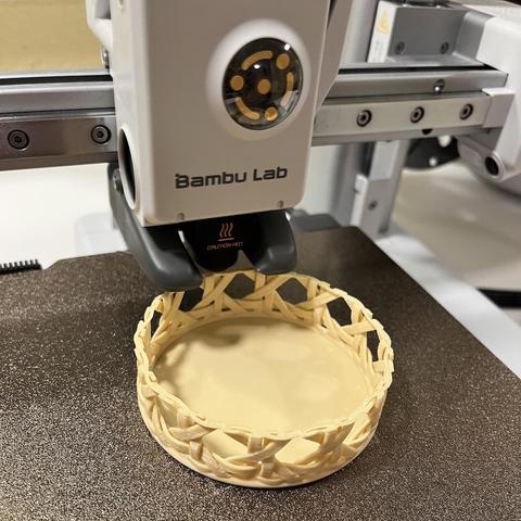 Photo showing the printbed of a BambuLab A1 Mini 3D printer, printing a circular basket with vienna canework weave in bambus like colored PLA.