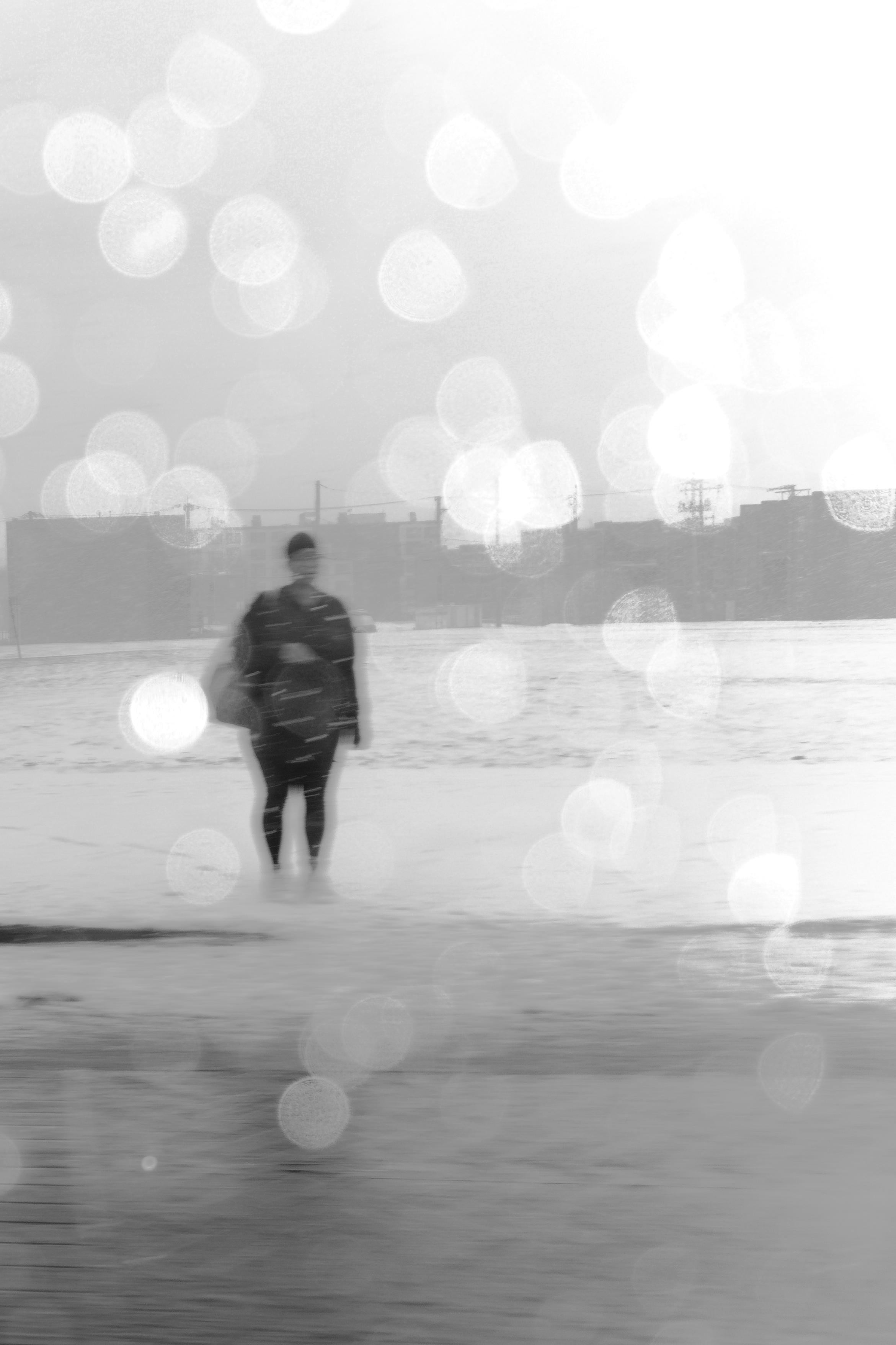 Black and white photograph of a woman waiting for a bus on an empty sidewalk. The window of the car I'm shooting through is snow-covered, creating little circular white flares throughout the image (about 2x the size of her head). The street in front of her is mostly featureless. Likewise, the empty lot behind her. Her clothing appears nearly black against an otherwise almost monochrome white image. Little snow streaks move right-to-left across her clothing.