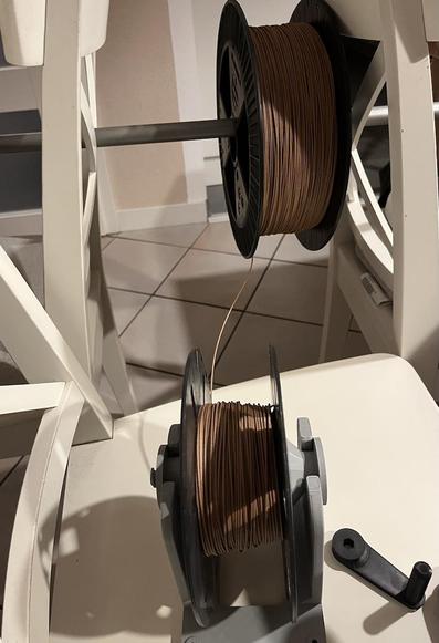The image shows a pair of chairs with a huge filament spool hanging on a broomstick and a normal 1 kg spool for a hobby 3D printer on another chair, with the first few windings already on the small spool. 
